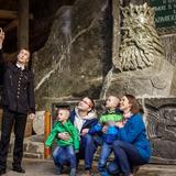 Zwiedzanie rodzinne w kopalni soli w Wieliczce. Czteroosobowa rodzina kuca pod solnym popiersiem króla Kazimierza Wielkiego i pokazuje dzieciom coś na stropie kopalni. Obok nich stoi przewodnik ubrany w czarny uniform.