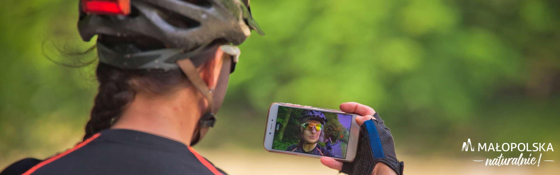 Kobietę, którą w ręku trzyma telefon z włączona aplikacją VisitMałopolska. W ekranie telefonu odbija się jej twarz. Ubrana jest w kas i okulary przeciwsłoneczne, na rękach ma rękawiczki do jazdy na rowerze.