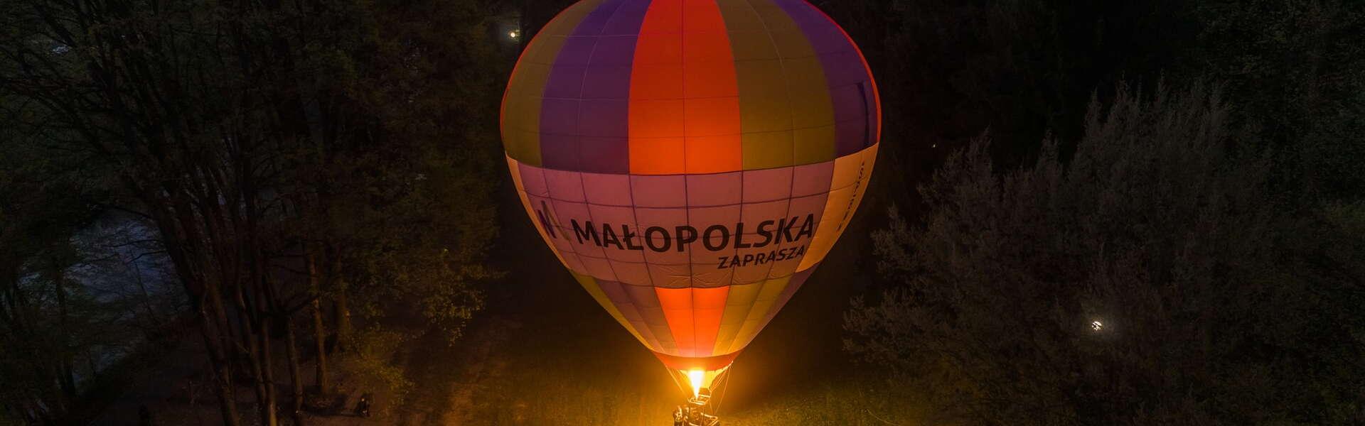 Balon na ogrzane powietrze z logo Małopolska w porze wieczornej w parku, podświetlony od dołu zapalonym palnikiem