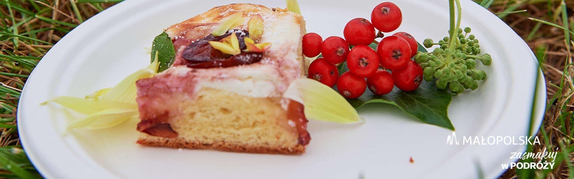 A piece of yeast cake with plums on a plate, next to the cake there is a decoration of rowan berries, in the lower right corner there is the Małopolska logo and the inscription 