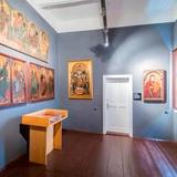 Sala muzealna, na niebieskich ścianach wiszą obrazy przedstawiające świętych, w centralnym punkcie białe drewniane drzwi.