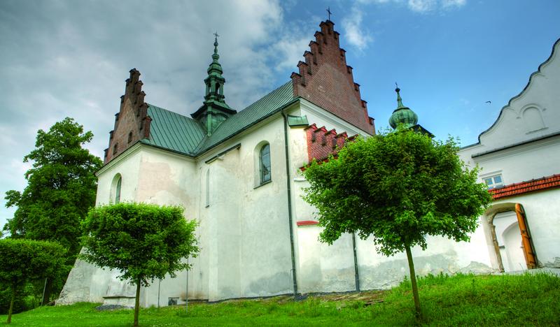 Kościół o białych ścianach z zielonymi dachami i brązowymi szczytami, z przodu drzewa o kulistych koronach.