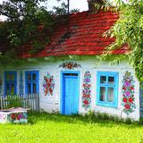 Na trawie stoi biała, drewniana chata z niebieskimi drzwiami i oknami, między którymi namalowane są bukiety kwiatów, z dachem pokrytym czerwoną dachówką i z kominem. Przed chatą, pomalowana w kwiaty kwadratowa studnia, przykryta blachą, za nią drewniany płotek ze sztachet. Po bokach rośną drzewa liściaste, które częściowo zakrywają zadaszenie.