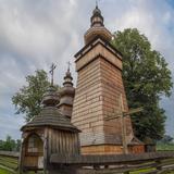 Na wprost z bliska wysoka, kwadratowa wieża z pochyłymi ścianami i z izbicą z koronką. Poniżej wysoki drewniany krzyż. Po lewej drewniana bramka z dachem gontowym i baniastym hełmem z gontu i krzyżem u góry. Z tyłu fragment dwóch dachów, jeden nad drugim nawy z baniastym hełmem i wieżyczką na sygnaturkę zwieńczoną u góry krzyżem. Z tyłu wysokie drzewa i zachmurzone niebo.