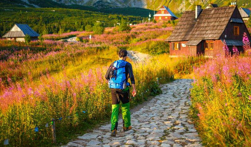 Kobieta idąca po kamiennym szlaku na Hali Gąsienicowej w Tatrach. Na końcu ścieżki stoi drewniana chata. Wokół jest dużo zieleni a trochę dalej Tatry.