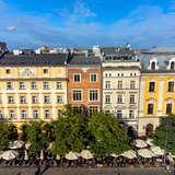 Rząd kolorowych kamienic na Rynku Głównym w Krakowie. Środkowa z nich to Kamienica pod Orłem. Przed budynkami znajduje się rząd parasoli oraz drzew.