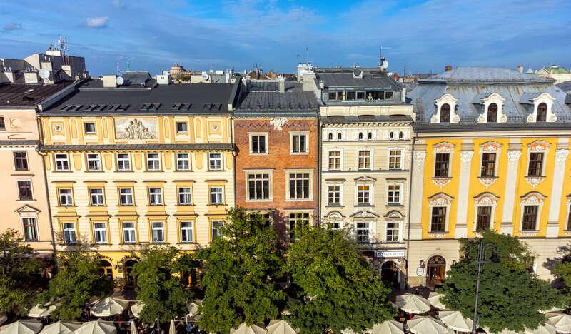 Rząd kolorowych kamienic na Rynku Głównym w Krakowie. Środkowa z nich to Kamienica pod Orłem. Przed budynkami znajduje się rząd parasoli oraz drzew.