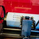 Maszyna drukarska na czerwonym tle, z dużym kołem i białą kartką papieru na której drukowane są nuty na pięcioliniach.