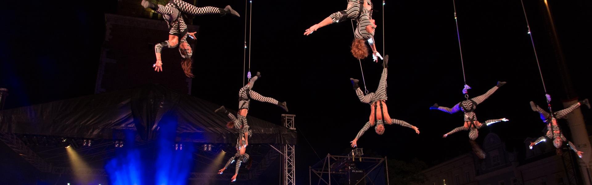 Kostümierte Schauspieler hängen kopfüber an Seilen über einer Menschenmenge während eines Theaterfestivals. Es ist dunkel, daneben gibt es eine blau beleuchtete Bühne.