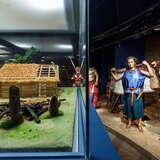 Po lewej stronie eskpozycji w Muzeum Archeologicznym w Krakowie znajduje się szklana gablota z inscenizacją wioski. Po prawej ludzkie postacie.