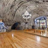 Widok na podziemną kaplicę świętej Kingi w Kopalni soli Bochnia. Z lewej krzyż wiszący na ścianie, po prawej ołtarz a przed nim ławki. Po środku kryształy żyrandol.