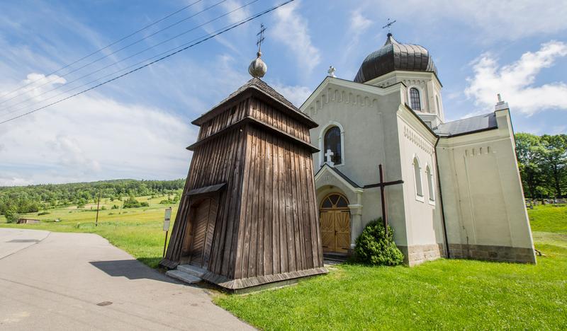 Drewniana dzwonnica stojąca przed murowanym, pomalowanym na jasny kolor budynkiem cerkwi.