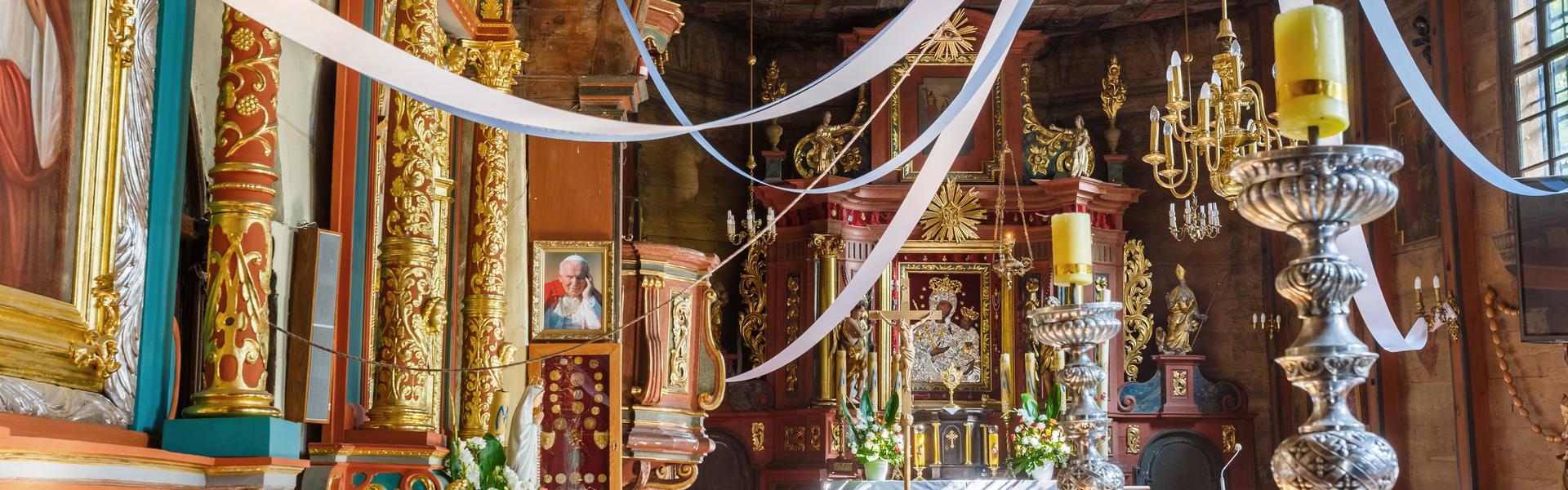 Wnętrze kościoła. Ołtarz główny w kolorze czerwieni i złota z obrazem Matki Boskiej. Po lewej stronie ambona. Dużo kwiatów, kościół jest przystrojony białymi i błękitnymi wstążkami, które z sufitu rozchodzą się promieniście po nawie.