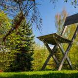 Pogodne letnie niebo, na tle drzew, na trawie stoi 14-metrowe drewniane, składane krzesło z oparciem.
