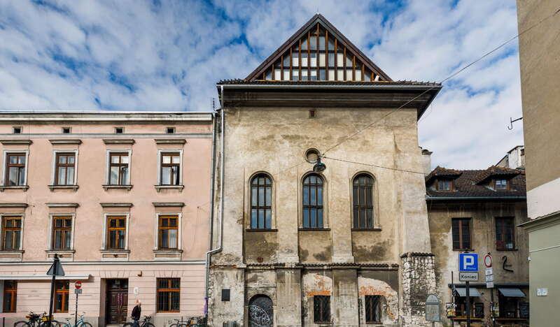 Stary, ciemnoszary budynek Synagogi Wysokiej w Krakowie, Z obu jej stron znajdują się kamienice, a pod budynkami widać rowery. Nad nimi widać niebieskie niebo z lekkimi chmurami.