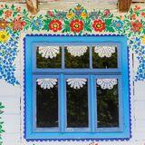 Okno drewnianego domu w niebieskich ramach, składające się z trzech części z mniejszymi okienkami u góry, ozdobione zazdrostkami w kształcie okrągłych serwetek, malowane naokoło w zalipiańskie kwiaty jakby lustrzane odbicie.