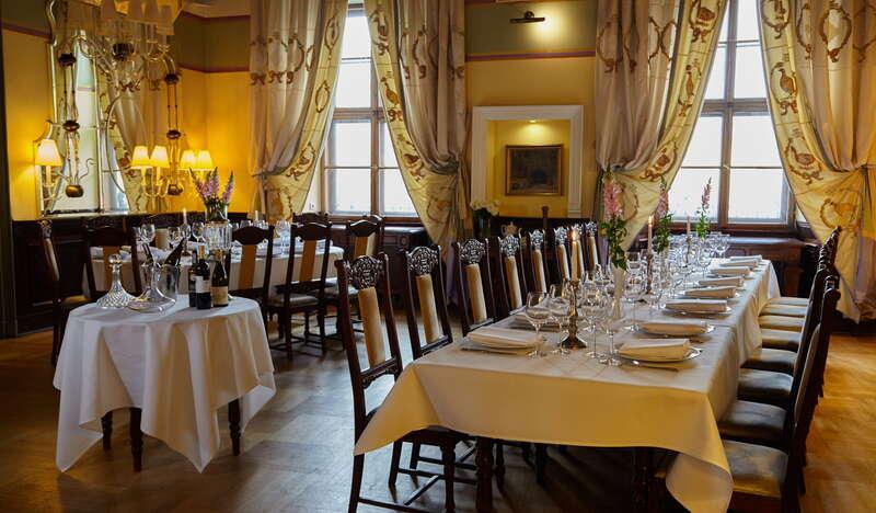 Dwa podłużne stoły na sali w Restauracji Wentzl w Krakowie. Oba nakryte białymi obrusami oraz przygotowane zastawą i kwiatami. Ściany sali są żółte a na oknach wiszą ozdobne zasłony.