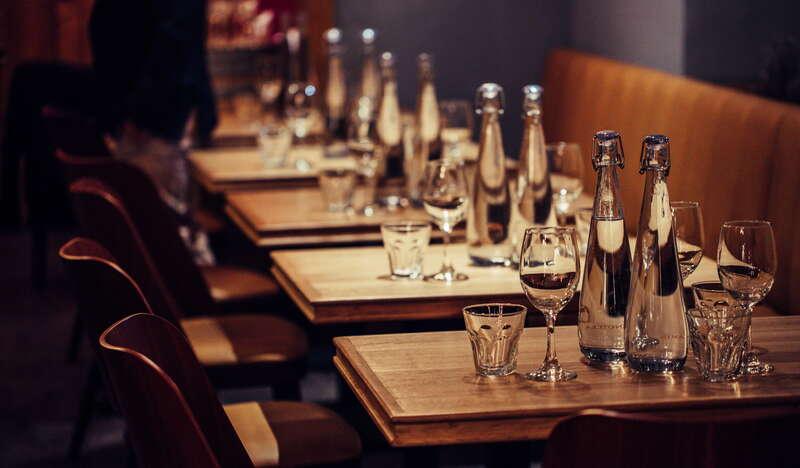 Drewniane stoliki w rzędzie w Restauracji Enoteka Pergamin w Krakowie. Na każdym stole znajduje się woda, szklanki oraz kieliszki. W oddali widać przechodzącego człowieka.