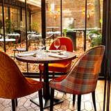 Trzy kolorowe krzesła przy stoliku w Restauracji Fiorentina w Krakowie. Za stołem znajdują się okna, a przez nie widać ogródek z krzesłami i stolikami, a także jesienne liście na ziemi.