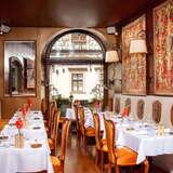 Dwa rzędy miejsc siedzących na górnej sali w Restauracji Pod Nosem w Krakowie. Na ścianach wiszą dywany, po lewej stronie na stoliku stoi alkohol, a na wprost znajduje się duże okno z widokiem na kamienicę.