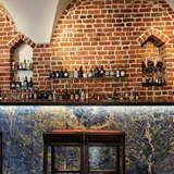 Widok na bar w Restauracji Trzy Rybki w Krakowie. Przy niebieskim, marmurowym barze stoi kilka barowych krzeseł. Nad barem, na ceglanej ścianie, na półkach stoją różnego rodzaju alkohole.