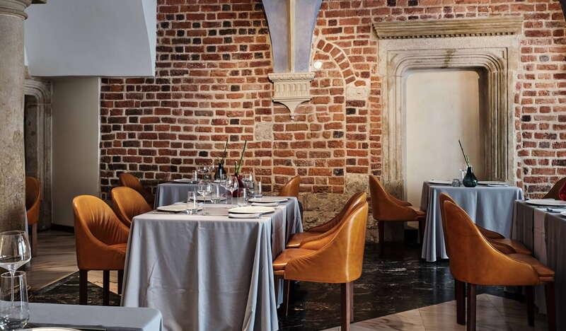 Widok na stoliki w Restauracji Trzy Rybki w Krakowie na tle ceglanej ściany. Przy stołach nakrytych szarym obrusem stoją żółte skórzane krzesła.