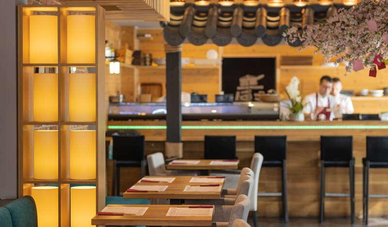 Sala w Restauracji Hana Sushi w Krakowie. Po lewej stronie znajduje się rząd stolików. Z tyłu widać kuchnię oraz dwóch kucharzy. Po prawej zwisają kwiaty z karteczkami.