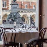 Widok na Sukiennice i pomnik Adama Mickiewicza z okna Restauracji Szarej w Krakowie. Okno znajduje się za okrągłym stolikiem z krzesłami.