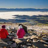Szczyt Babia Góra. Na pierwszym planie dwie osoby siedzące tyłem do obiekty na kamieniu.  W oddali widać mgłę