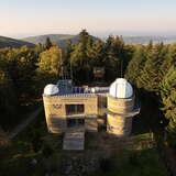 Bryłę budynku obserwatorium astronomicznego na szczycie Lubomir. Budynek  z dwoma kopułami i płaskim dachem. Wokół drzewa.