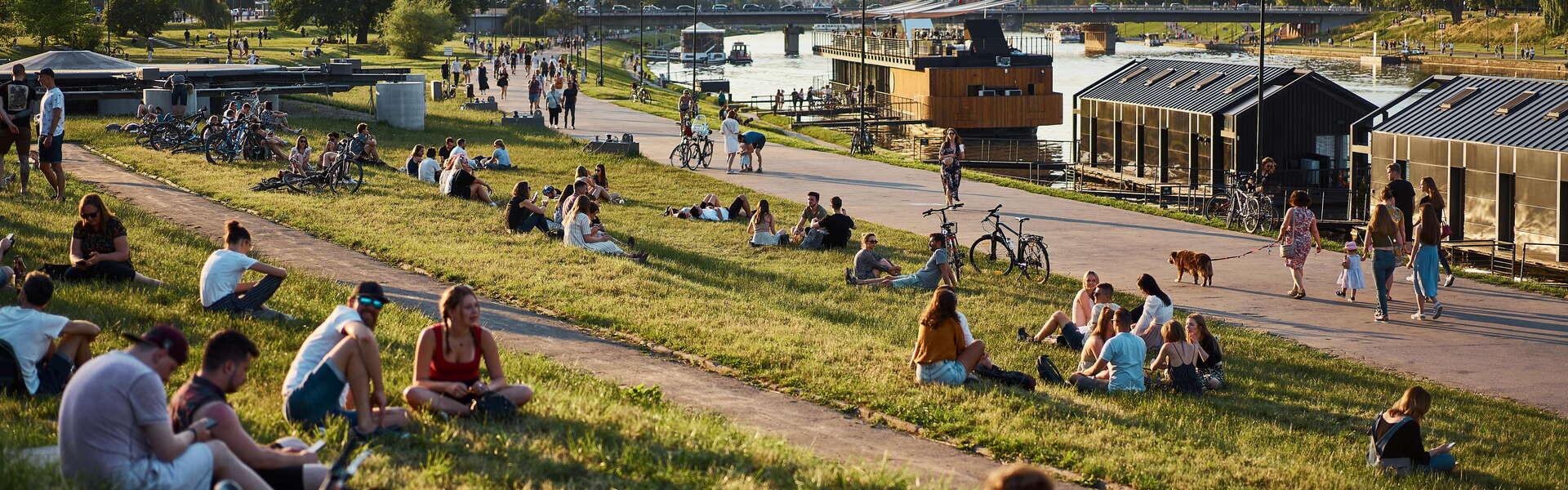 Blick auf Gruppen von Menschen, die auf dem Rasen an der Weichsel-Promenade in Kraków sitzen. Rechts am Ufer liegen Schiffe vor Anker. Im Hintergrund ein Fluss und eine lange Brücke. Weiter links Bäume, und rechts auf dem anderen Ufer das Königsschloss Wawel mit zwei runden Türmen. Klarer sonniger Himmel.