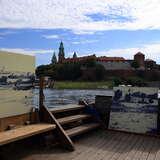 Fragment drewnianej łodzi na rzece Wiśle, na której stoją obrazy rysowane ołówkiem, przedstawiające wzgórze Wawel z Zamkiem Królewskim i dwoma wieżami Katedry na Wawelu.