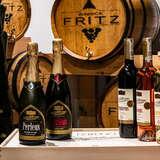 Butelki z winem produkcji Winnicy Fritz stojące na drewnianej skrzyni. Za nimi znajdują się drewniane beczki, a obok na regale postawione szklane butelki z winem.