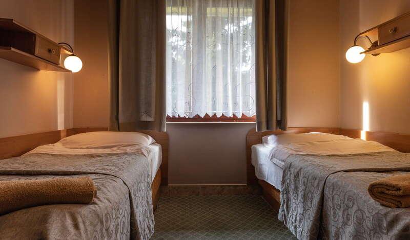 Pokój hotelowy, dwa pojedyncze łóżka pod ścianami, lampki, okno z firanką.