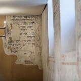 Wnętrze synagogi Kupa w Krakowie. Przybliżenie na ścianę z napisami po hebrajsku. Obok ściana również z napisami i malunkami, w niej trzy otwory z oknami. U sufitu wisi metalowy żyrandol z żarówkami w formie świec.