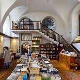Długi stół zastawiony książkami w sklepie w Synagodze Poppera w Krakowie. Z tyłu przy ścianie widać drewniane schody, a przy lewej ścianie stoi kobieta w płaszczu,