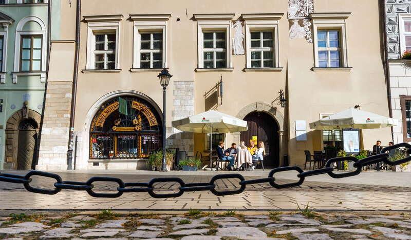 Kawałek wybrukowanej ulicy i łańcuch, a zaraz nad nią Kamienica Hipolitów w Krakowie. Przed budynkiem stoją dwie parasolki, widać też ludzi oraz sklep z pamiątkami we wnętrzu.