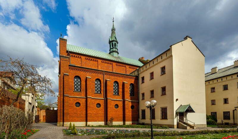 Kościół pw. Niepokalanego Serca Maryi w Krakowie z zewnątrz. Widać dwie części świątyni, jedna ceglana, a druga gładka beżowa. Budynek jest otoczony murem, a koło niego znajduje się ogród z kwiatami.