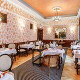 Pomieszczenie restauracji z ustawionymi stolikami i krzesłami. Na ścianie tapeta. U sufitu zwisające eleganckie stylizowane żyrandole.