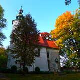 Biały, kamienny niewielki kościół z wieżą od frontu zakończoną hełmem z sygnaturką, pokryty czerwonym dwuspadowym dachem. Wokół kilka drzew w kolorach jesieni.