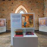 Ekspozycja muzealna z tablicą, na której opisane są dzieje o cechach rzemieślniczych w Krakowie w XIV wieku. Na pierwszym planie szklana gablota za którą znajduje się drewniana skrzynka. W tle obrazy postaci. Po lewej drewniana, przeszklona gablota.