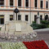 Pomnik - wzniesienie z kamieni zwieńczone czarną metalową urną. Przed pomnikiem biało-czerwone kwiaty. Z tyłu budynek, po prawej stronie pomnik.