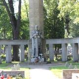 Fragment kamiennego obelisku otoczonego półokrągłą kolumnadą z daszkiem. Przed obeliskiem stoi monumentalna wysoka figura rycerza-strażnika wspartego na mieczu. Z tyłu drzewa.