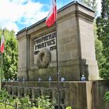 Duży kamienny pomnik z łacińskim napisem Pro Patria Mortuis 1914–1918. Po jego dwóch stronach flagi Polski, za nim drzewa.